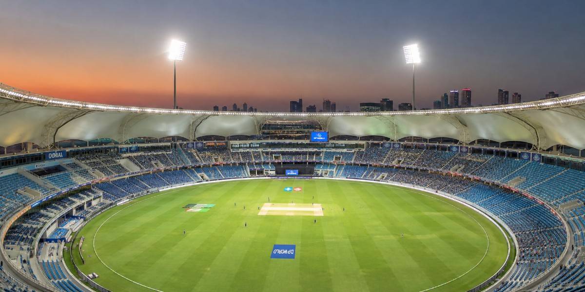 Dubai International Stadium panoramic view with the cricket pitch, blue seating, floodlights, and city skyline at dusk.